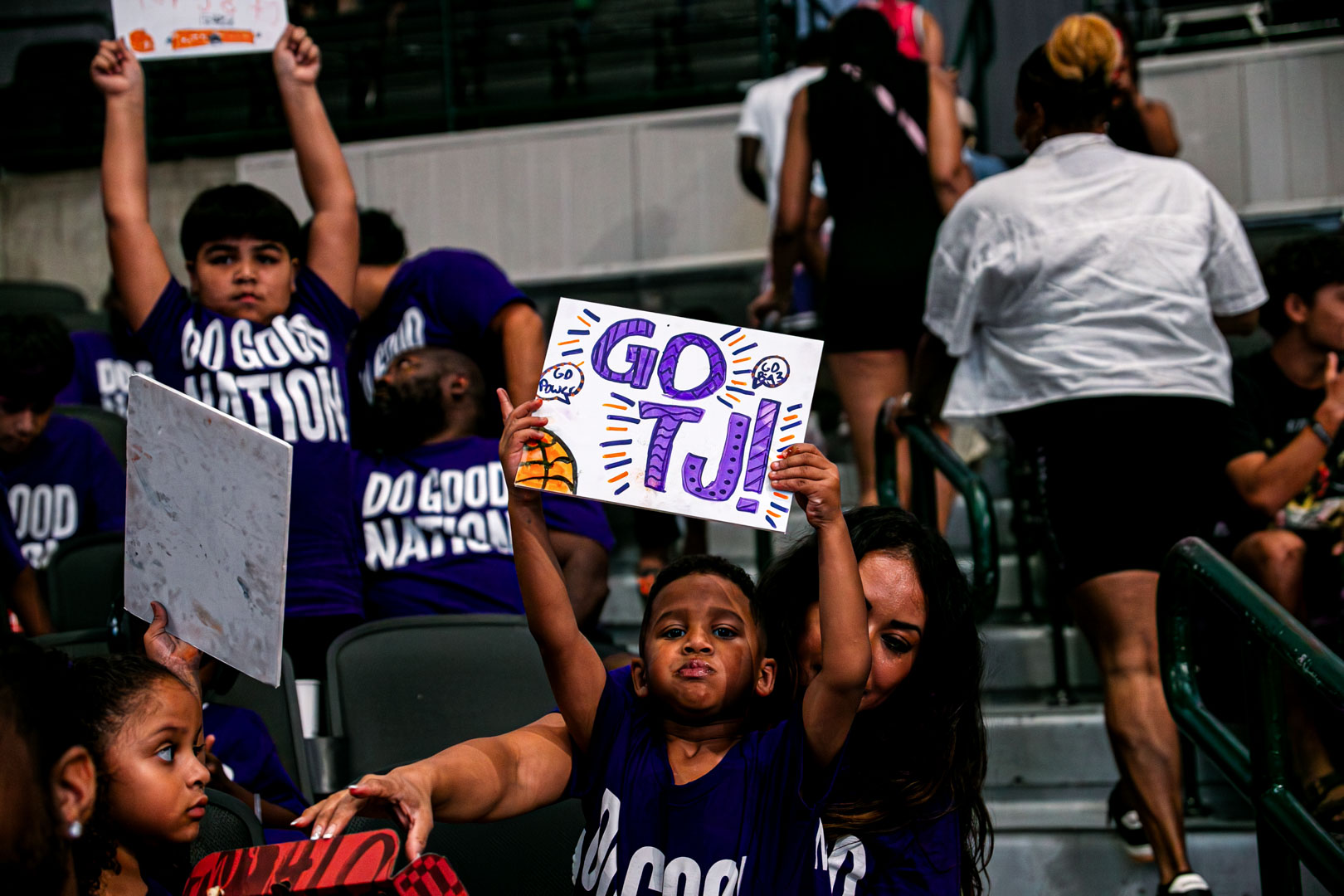 Youth in purple Do Good Nation shirts in the stands, one boy holding a handmade "GO TJ!" sign above his head