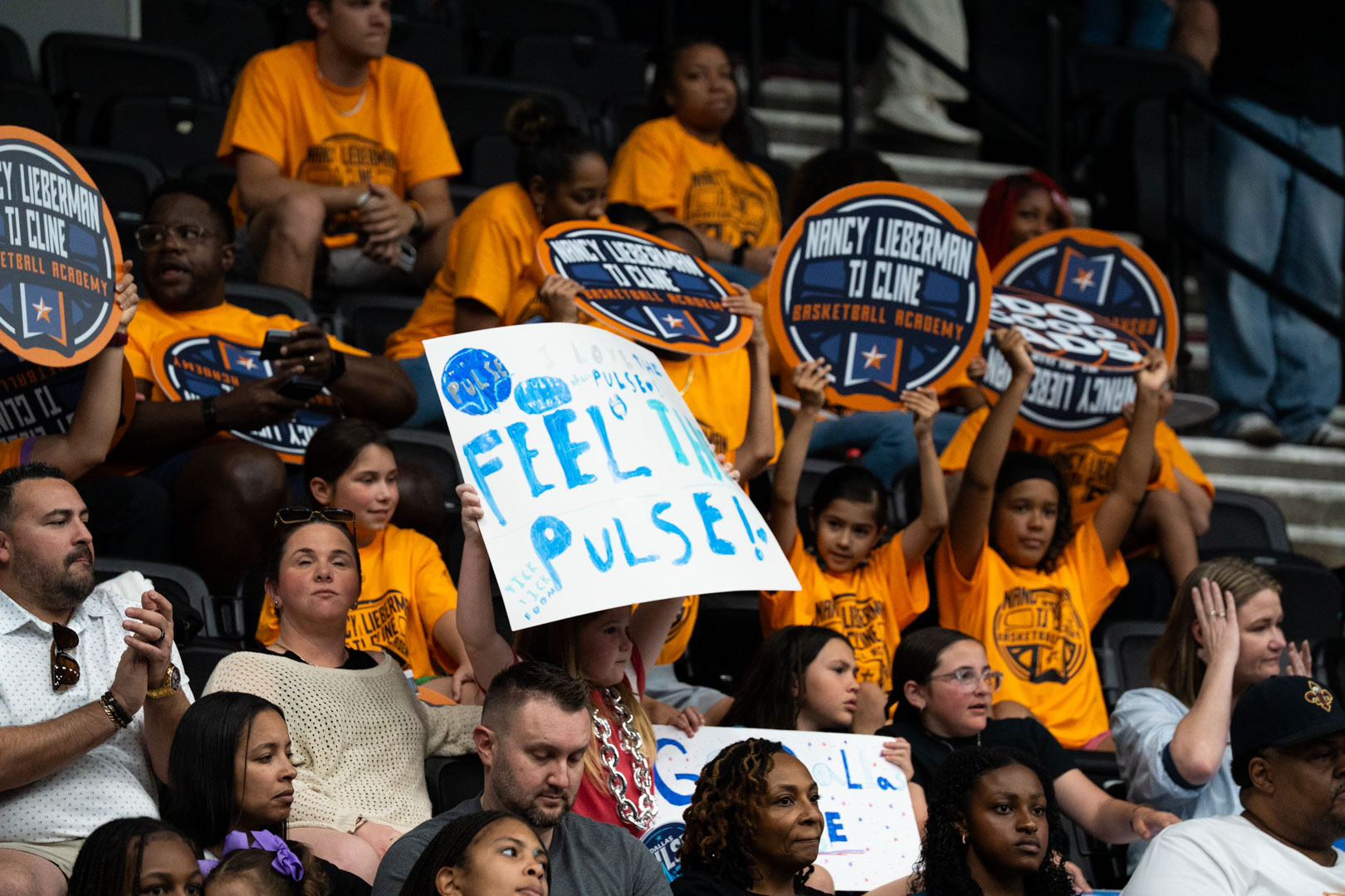 Youth in matching orange Nancy Lieberman TJ Cline Basketball Academy shirts filling the stands, holding team paddles and a handmade "Feel the Pulse!" sign