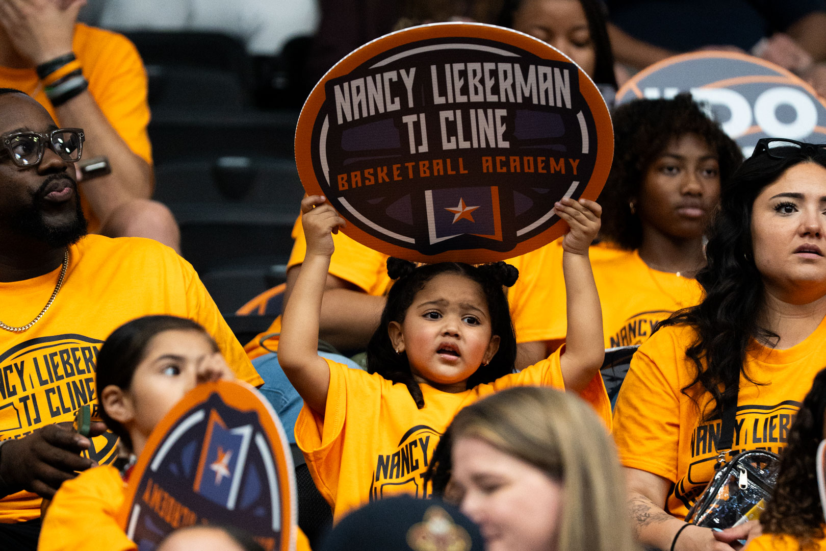A young girl holding a Nancy Lieberman TJ Cline Basketball Academy paddle above her head in the stands